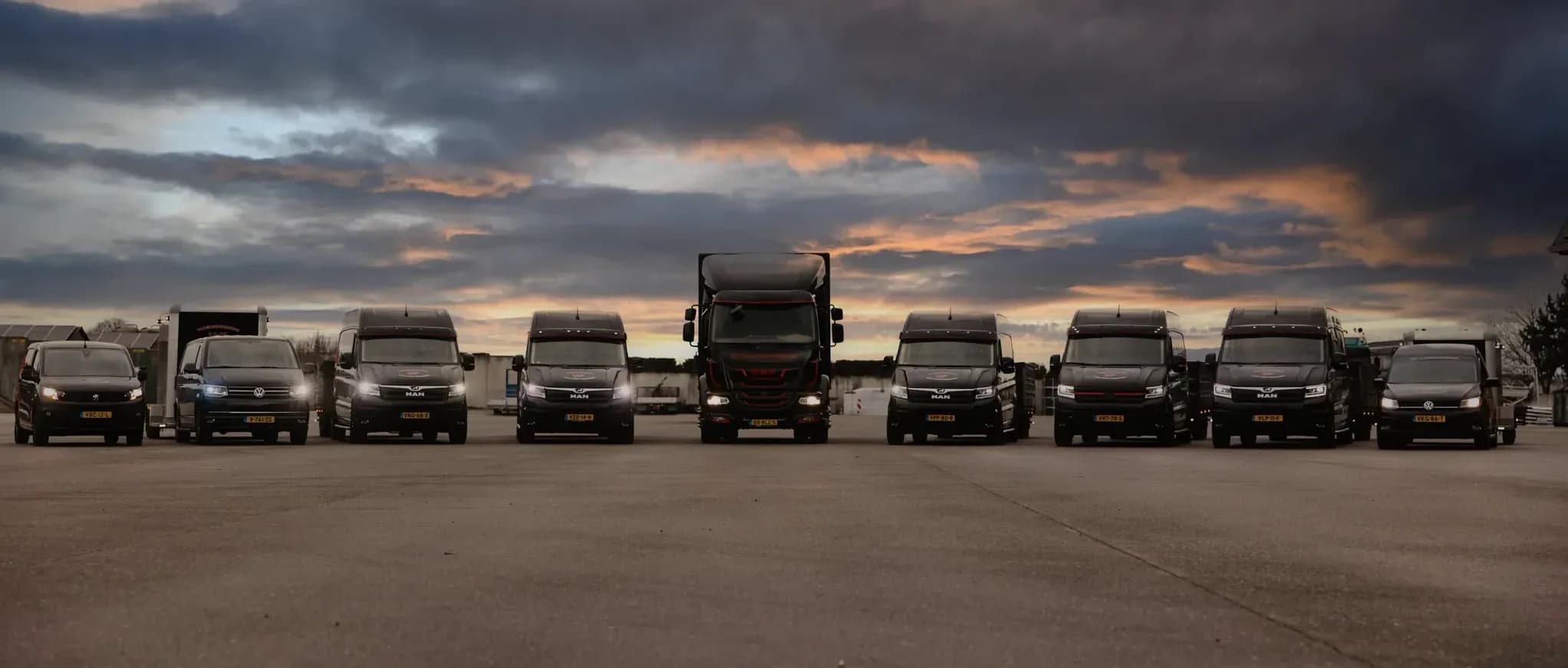 Rij geparkeerde bestelwagens en vrachtwagens op een parkeerterrein met een kleurrijke, bewolkte avondlucht