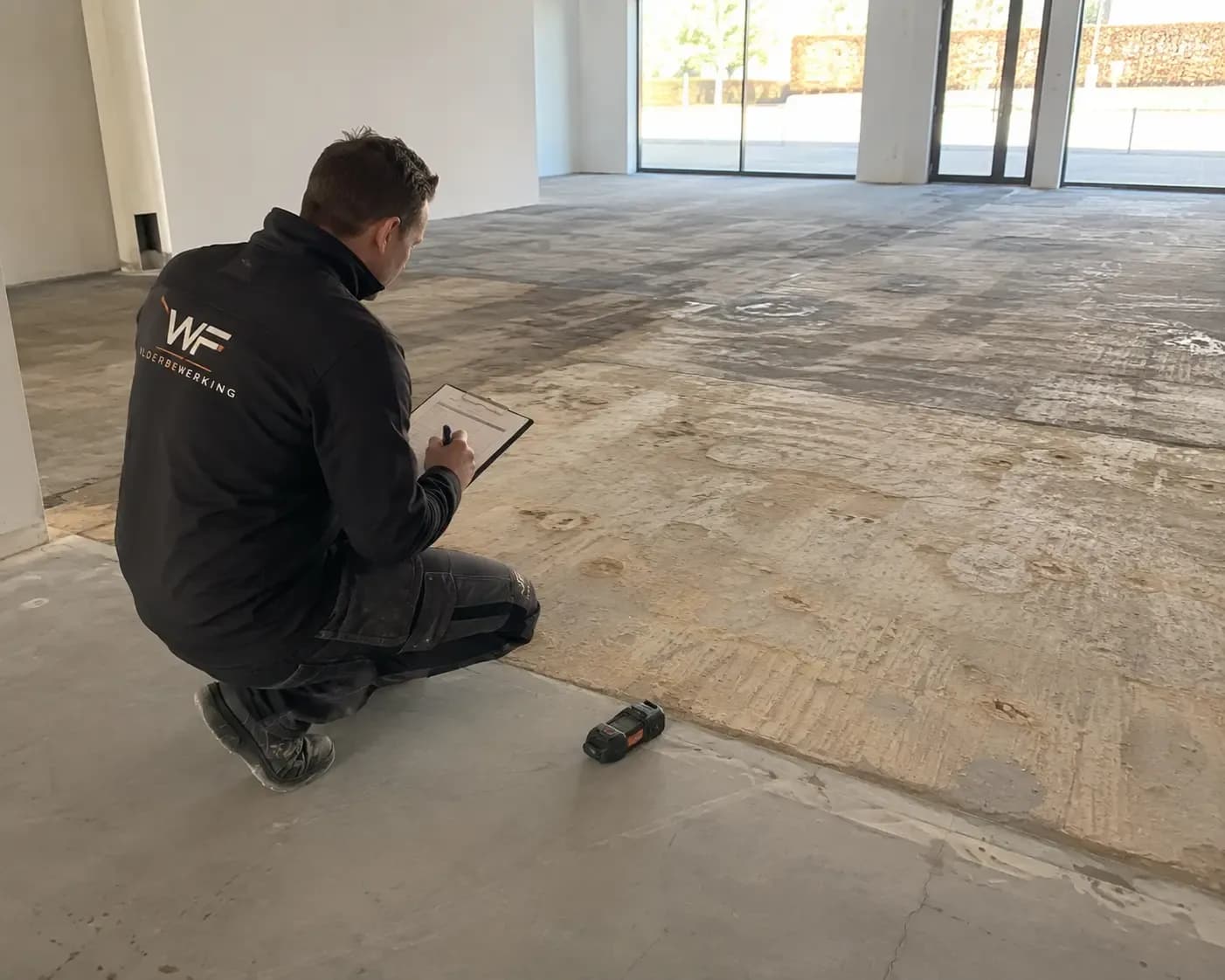 Werker in werkkleding knielt en vult formulier op schrijfplank tijdens inspectie van een betonvloer met resten van verwijderde vloerbedekking; jas toont letters 'WF'.